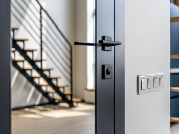 Close view of a switch block beside a black framed glass door handle, metal stairs and soft shadows in the background create a modern loft minimal vibe.